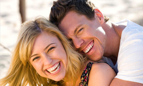 Picture of a smiling couple, happy with the prices they received for dental work at the Costa Rica Dental Center in San Jose, Costa Rica.  The woman has sandy blonde hair, the man has short dark hair and both are hugging and showing their perfect teeth while smiling directly at the camera.